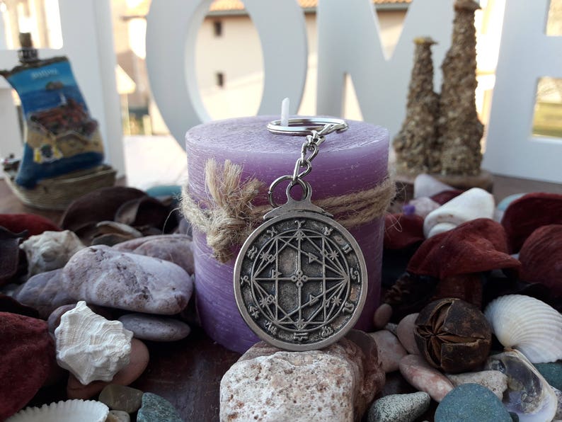 May include: A silver charm with a geometric design hangs from a silver key ring. The charm is in front of a purple candle with a brown twine bow.