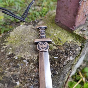 May include: A silver-colored miniature sword with a double-headed eagle design on the pommel. The sword is lying on a rough, gray surface with green moss.