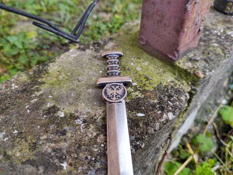 May include: A silver-colored miniature sword with a double-headed eagle design on the pommel. The sword is lying on a rough, gray surface with green moss.