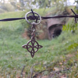 Puede incluir: Un llavero con colgante de nudo celta plateado. El colgante cuelga de un anillo de metal sobre un fondo borroso de follaje verde y una entrada de t&uacute;nel. Un accesorio decorativo.