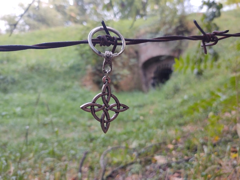 Puede incluir: Un llavero con colgante de nudo celta plateado. El colgante cuelga de un anillo de metal sobre un fondo borroso de follaje verde y una entrada de t&uacute;nel. Un accesorio decorativo.