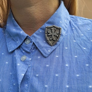 May include: A silver-toned metal pin shaped like a shield with a cross in the center. The pin is attached to a blue and white polka dot shirt.