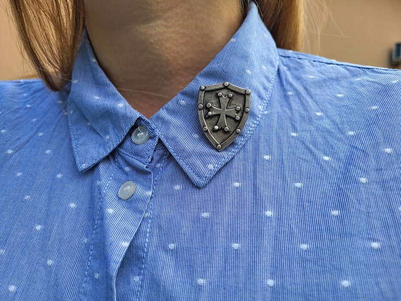 May include: A silver-toned metal pin shaped like a shield with a cross in the center. The pin is attached to a blue and white polka dot shirt.