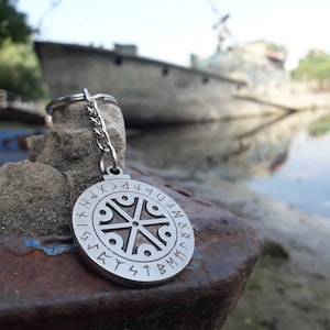 May include: A silver keychain with a circular pendant featuring a symbol surrounded by runes. The pendant is hanging from a silver chain.