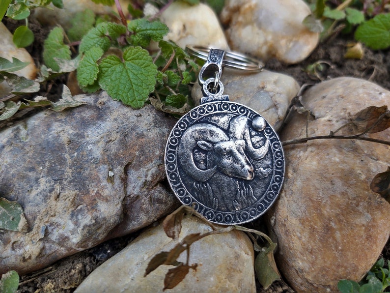 May include: Silver key chain with a round charm featuring a detailed ram head design. The charm is surrounded by a decorative border.