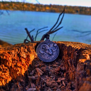 May include: A silver pendant featuring an intricate design of a ram's head. The pendant is positioned on a weathered tree stump with a blurred backdrop of a lake and trees.