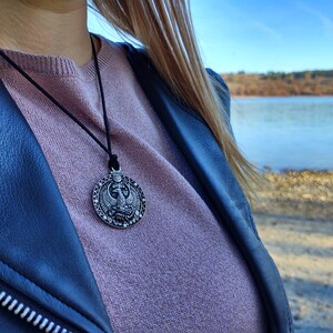 May include: A silver pendant necklace featuring an intricate design of a bird with outstretched wings. The pendant is round and hangs from a black leather cord.