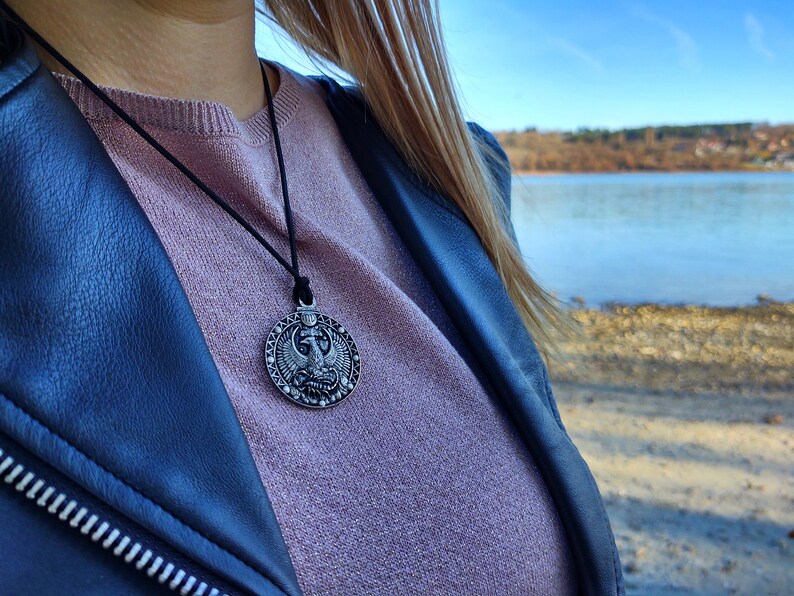 May include: A silver pendant necklace featuring an intricate design of a bird with outstretched wings. The pendant is round and hangs from a black leather cord.