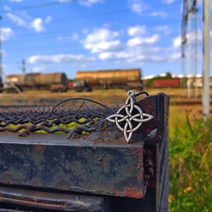 Puede incluir: Un colgante plateado con un nudo celta en un cord&oacute;n negro, apoyado sobre una superficie met&aacute;lica oxidada. El dise&ntilde;o intrincado del colgante est&aacute; enfocado, con un fondo borroso de vagones de tren y cielo azul.