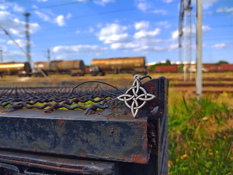 Puede incluir: Un colgante plateado con un nudo celta en un cord&oacute;n negro, apoyado sobre una superficie met&aacute;lica oxidada. El dise&ntilde;o intrincado del colgante est&aacute; enfocado, con un fondo borroso de vagones de tren y cielo azul.