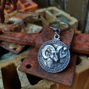May include: Silver-toned keyring with a circular charm featuring a detailed ram's head design. The charm is surrounded by a decorative border of circles. The keyring is resting on a pile of rusty metal objects.