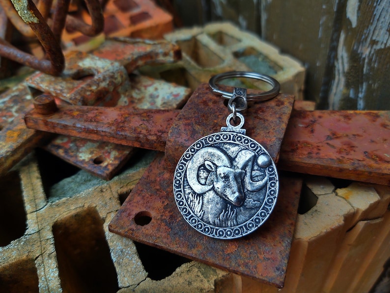 May include: Silver-toned keyring with a circular charm featuring a detailed ram's head design. The charm is surrounded by a decorative border of circles. The keyring is resting on a pile of rusty metal objects.