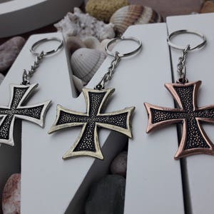 Puede incluir: Tres llaveros plateados con cruces de diferentes colores. Las cruces son plateadas, doradas y cobrizas. Cada cruz tiene una superficie texturizada y un diseño detallado.