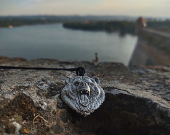 May include: A silver-toned metal pendant shaped like a roaring bear head with detailed fur texture and visible teeth, suspended from a thin black cord. The pendant features a three-dimensional design with dark oxidized accents in the recessed areas.