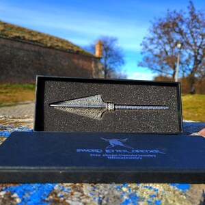 May include: A silver-colored arrowhead-shaped knife with a textured surface is displayed in a black box with a black foam interior. The box is open and the knife is visible. The box has the text "SWORD ETTER OFIXER" and "Etsy shop: Danubejewelry" printed on it.