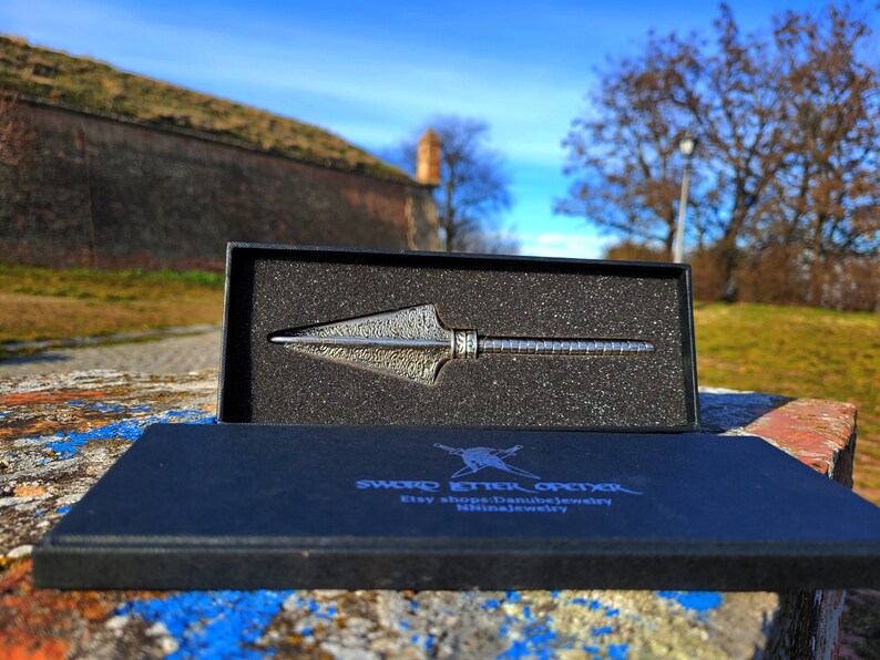 May include: A silver-colored arrowhead-shaped knife with a textured surface is displayed in a black box with a black foam interior. The box is open and the knife is visible. The box has the text "SWORD ETTER OFIXER" and "Etsy shop: Danubejewelry" printed on it.