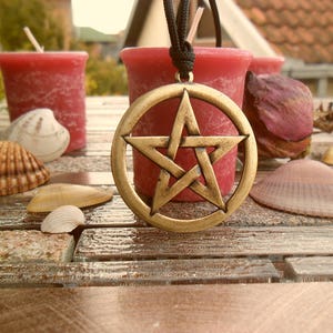 May include: A gold pentagram pendant necklace on a black cord, hanging over a red candle. The pendant is surrounded by seashells and a rose on a wooden surface.