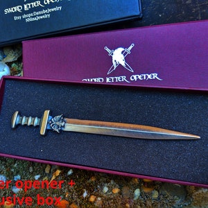 Puede incluir: Un abrecartas con forma de espada de color bronce, con un mango decorativo, presentado en una caja burdeos. La caja lleva la inscripci&oacute;n "SWORD LETTER OPENER". El abrecartas se muestra en un inserto de espuma negra.
