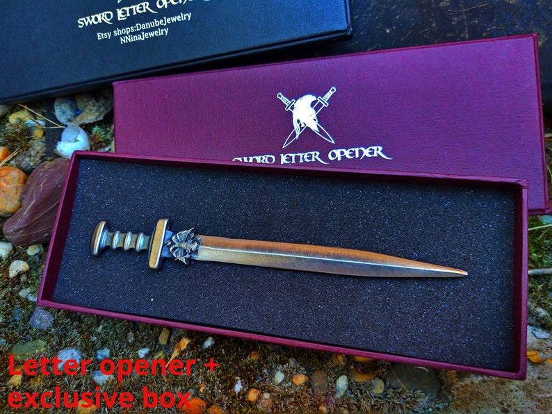 Puede incluir: Un abrecartas con forma de espada de color bronce, con un mango decorativo, presentado en una caja burdeos. La caja lleva la inscripci&oacute;n "SWORD LETTER OPENER". El abrecartas se muestra en un inserto de espuma negra.