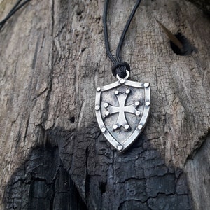 May include: A silver shield pendant necklace with a cross design. The pendant is hanging from a black cord.