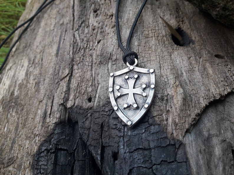 May include: A silver shield pendant necklace with a cross design. The pendant is hanging from a black cord.