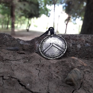 May include: A silver pendant necklace with a shield design and chevron detail. The round pendant has a textured finish and hangs from a black cord. The necklace is displayed against a natural, earthy backdrop.