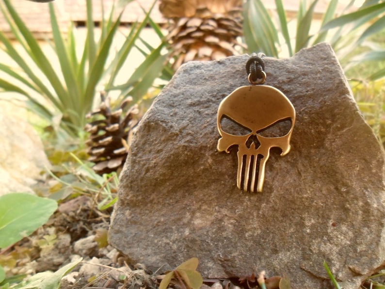 Pode incluir: Um colar com um pingente de caveira dourado e um cord&atilde;o preto. O pingente &eacute; uma representa&ccedil;&atilde;o detalhada de uma caveira com uma mand&iacute;bula proeminente e uma s&eacute;rie de linhas verticais na mand&iacute;bula inferior. O colar est&aacute; apoiado em uma rocha marrom e &aacute;spera.