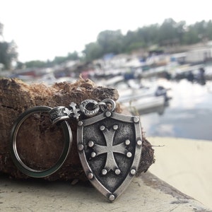 May include: A silver keychain with a shield-shaped charm featuring a cross and a ring. The charm is detailed with small dots around the edge and the cross.