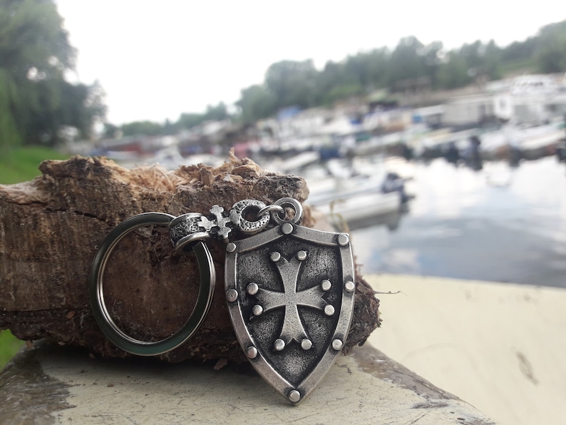 May include: A silver keychain with a shield-shaped charm featuring a cross and a ring. The charm is detailed with small dots around the edge and the cross.