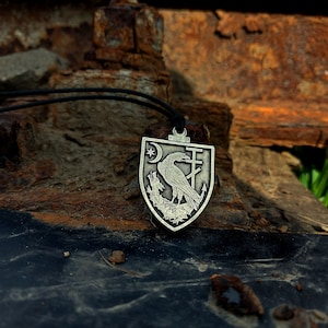 May include: A silver pendant necklace with a shield-shaped charm featuring a raven, a crescent moon, and a cross. The pendant is on a black cord and is resting on a rusty metal surface.