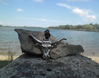 Może przedstawiać: Srebrny naszyjnik z zawieszką w kształcie głowy byka na czarnym sznurku. Zawieszka leży na skale w pobliżu zbiornika wodnego.