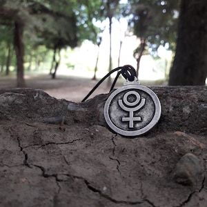 May include: A silver pendant necklace with a circular charm featuring the astrological symbol for Uranus. The pendant is on a black cord and is lying on a cracked, brown surface.