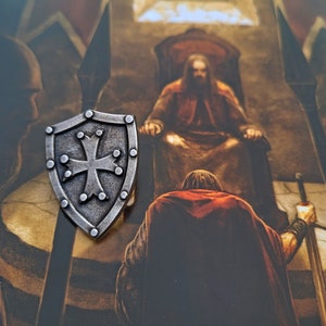 May include: A silver shield with a cross in the center, surrounded by small dots. The shield is on a brown background with a painting of a person in a throne.
