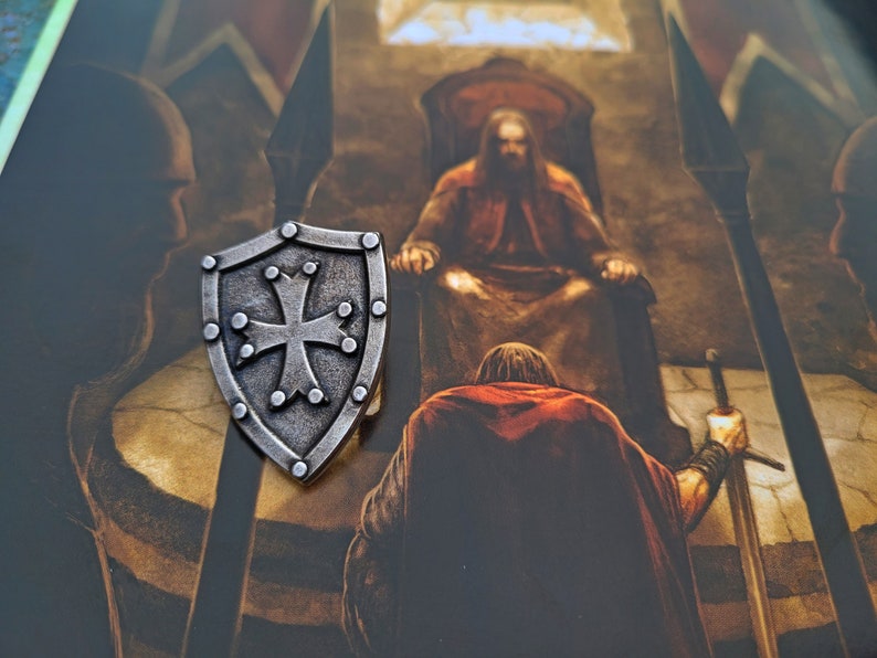 May include: A silver shield with a cross in the center, surrounded by small dots. The shield is on a brown background with a painting of a person in a throne.