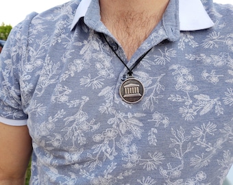 May include: A silver necklace with a round pendant featuring a stylized image of a building with columns. The necklace is worn by a person wearing a blue shirt with a white floral pattern.