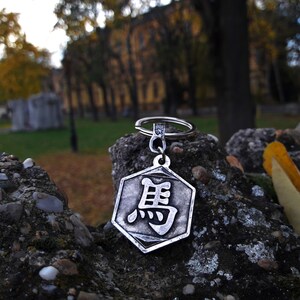 May include: A silver-colored keychain with a hexagonal charm featuring a Chinese character. The keychain is resting on a rock with a blurred background of trees and a building.