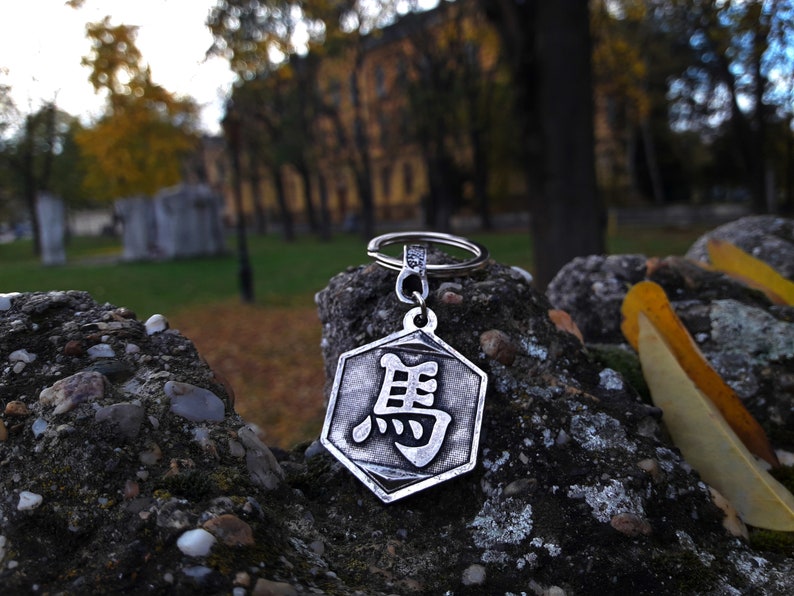 May include: A silver-colored keychain with a hexagonal charm featuring a Chinese character. The keychain is resting on a rock with a blurred background of trees and a building.
