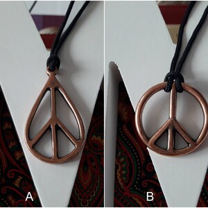 May include: Two necklaces with peace sign pendants. The pendant on the left is a teardrop shape with a peace sign inside. The pendant on the right is a round peace sign. Both pendants are copper colored and the necklaces are black.