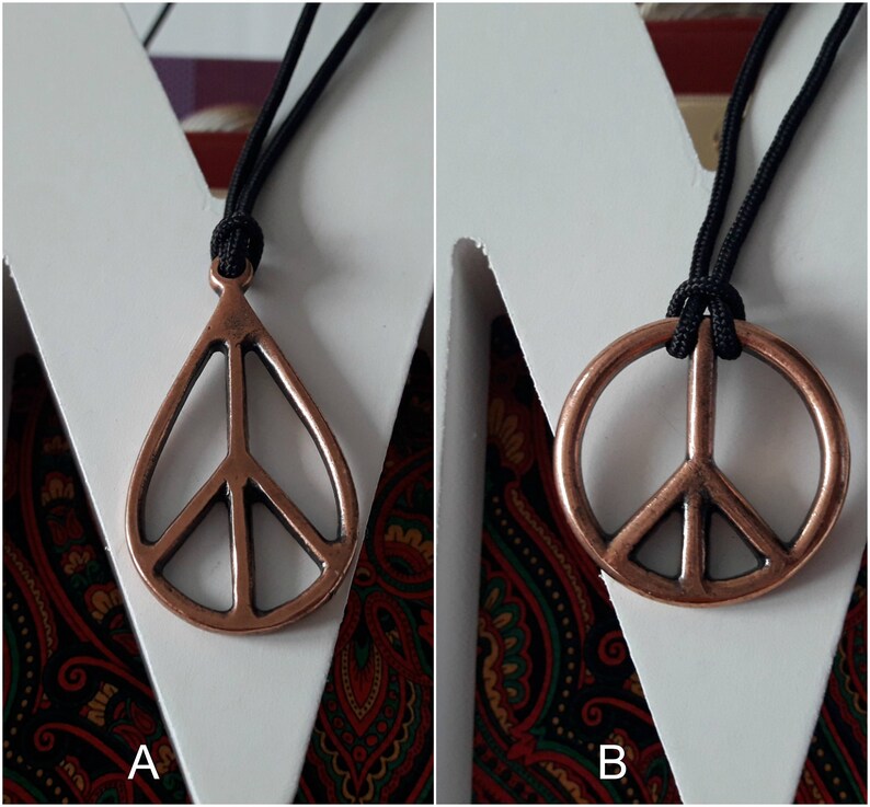 May include: Two necklaces with peace sign pendants. The pendant on the left is a teardrop shape with a peace sign inside. The pendant on the right is a round peace sign. Both pendants are copper colored and the necklaces are black.