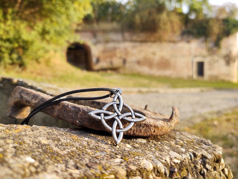 Puede incluir: Collar con colgante de nudo celta plateado y cord&oacute;n negro. El colgante presenta un dise&ntilde;o sim&eacute;trico y detallado. El collar descansa sobre un objeto met&aacute;lico oxidado y desgastado, con un fondo borroso de una estructura de piedra.