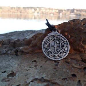 May include: A silver pendant with a circular design featuring a complex geometric pattern and symbols. The pendant is hanging from a black cord and is resting on a rock.