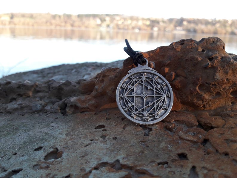 May include: A silver pendant with a circular design featuring a complex geometric pattern and symbols. The pendant is hanging from a black cord and is resting on a rock.