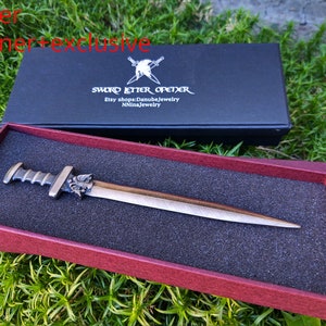 Puede incluir: Un abrecartas de color bronce con dise&ntilde;o de espada, presentado en una caja negra con el texto "SWORD LETTER OPENER". El abrecartas se muestra en una caja roja con un inserto de espuma negra.
