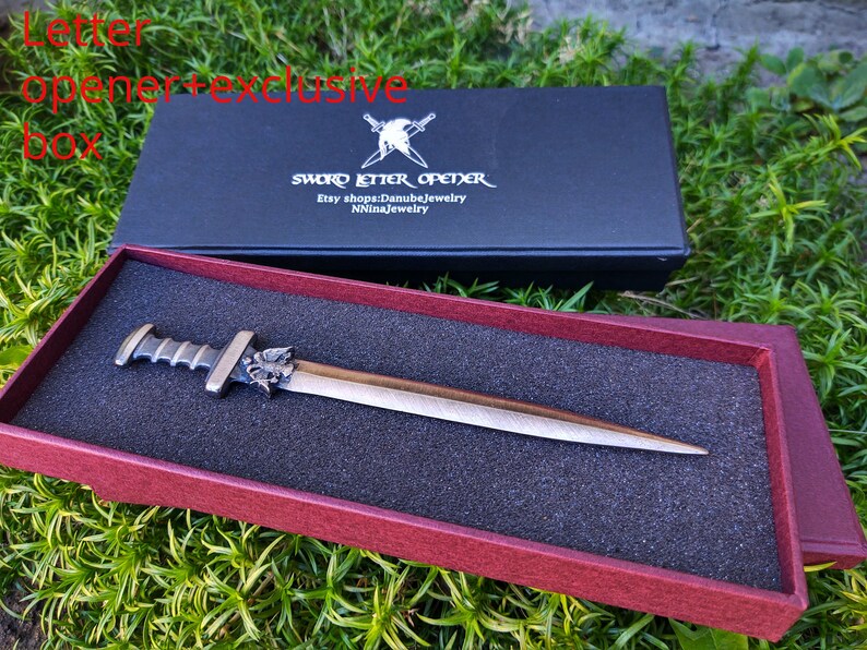 Puede incluir: Un abrecartas de color bronce con dise&ntilde;o de espada, presentado en una caja negra con el texto "SWORD LETTER OPENER". El abrecartas se muestra en una caja roja con un inserto de espuma negra.