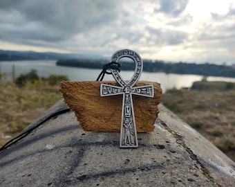 Pode incluir: Um pingente de Ankh de prata com hieróglifos descansando em um pedaço de madeira. O pingente está pendurado em um cordão preto. O fundo é uma imagem desfocada de um rio e um céu nublado.