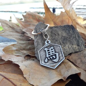 May include: A silver keychain with a hexagonal charm featuring a Chinese character. The charm is on a rock surrounded by brown leaves.
