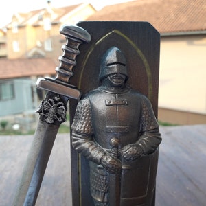 Puede incluir: Escultura detallada de un caballero con armadura, sosteniendo una espada. La espada tiene un mango decorativo y un emblema de &aacute;guila bic&eacute;fala. El caballero est&aacute; frente a un panel de madera oscura con borde dorado.