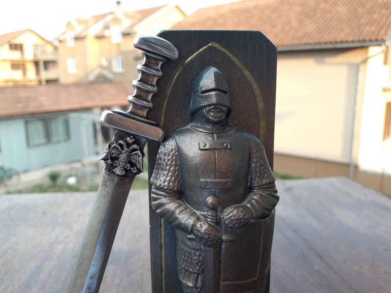 Puede incluir: Escultura detallada de un caballero con armadura, sosteniendo una espada. La espada tiene un mango decorativo y un emblema de &aacute;guila bic&eacute;fala. El caballero est&aacute; frente a un panel de madera oscura con borde dorado.