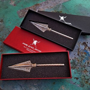 May include: Three boxes with a silver letter opener inside. The boxes are red, black, and black. The letter opener has a decorative, textured blade and a silver handle. The red box has the text "Sword Letter Opener" printed on it.