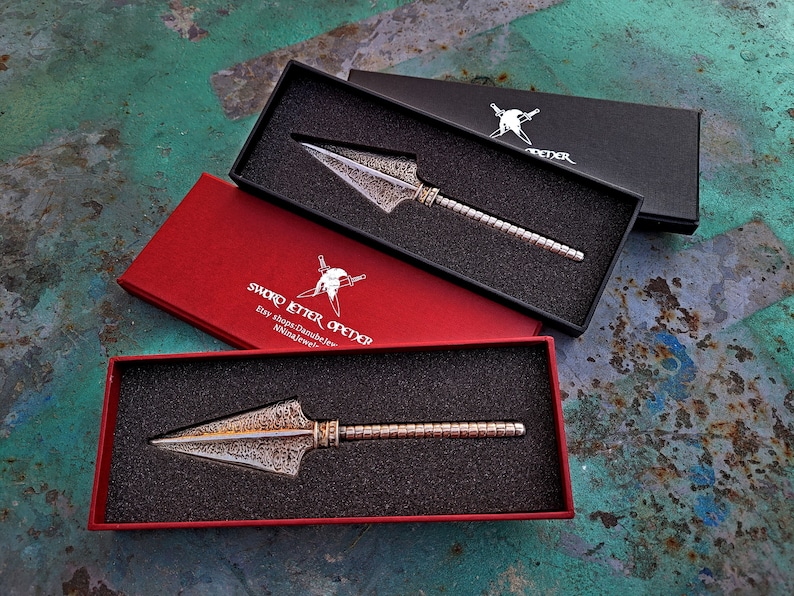 May include: Three boxes with a silver letter opener inside. The boxes are red, black, and black. The letter opener has a decorative, textured blade and a silver handle. The red box has the text "Sword Letter Opener" printed on it.